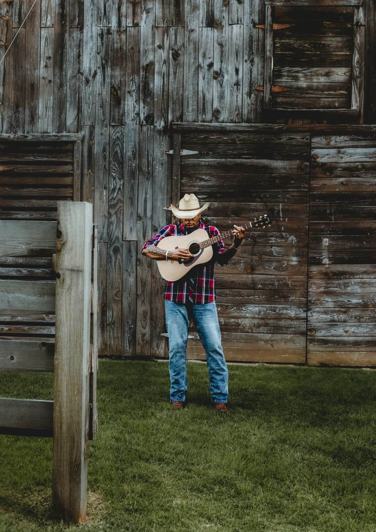 a man in jeans, boots, a western shirt, cowboy hat and guitars standing in front of a weathered wooden barn